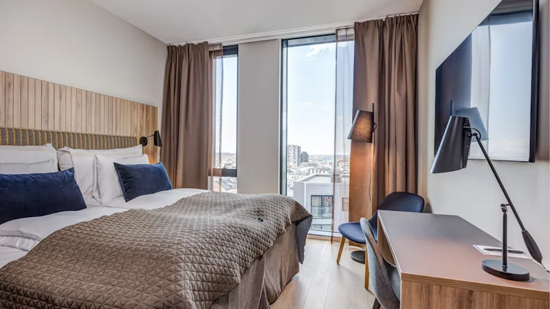 Et lyst og moderne hotelværelse med en hyggelig seng, et funktionelt arbejdsområde og store vinduer med udsigt over byens skyline.