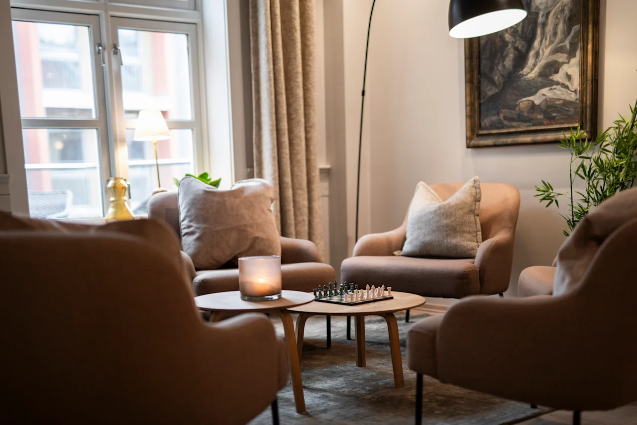 Fire beige lænestole omkranser et lavt bord med et skakspil og stearinlys, placeret i en hyggelig stue med et stort vindue, gardiner og kunst på væggen.