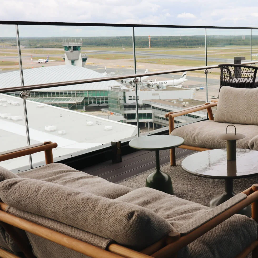 Outdoor seating area, with cushioned sofas and tables, overlooking an airport runway. Glass barriers surround the balcony, providing a view of planes and control tower in the distance.