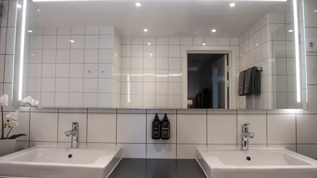 Large, illuminated mirror above two white sinks in a bathroom. Clean tiled walls and ceiling with small ceiling lights. Black soap dispensers and towels enhance the modern aesthetic.