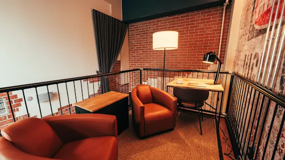 A cozy seating area with two orange armchairs, a small desk, and lamps, set against a brick wall and metal railing.
