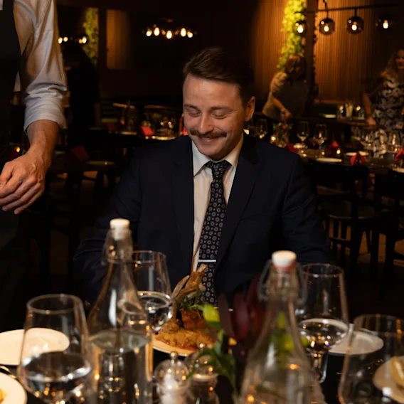 En person i dress smiler mens en kelner serverer et måltid ved et restaurantbord, omgitt av dekketøy.
