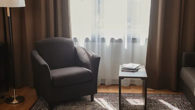 En grå fåtölj står bredvid ett litet bord med böcker, i ett soligt rum med gardiner och en mönstrad matta.