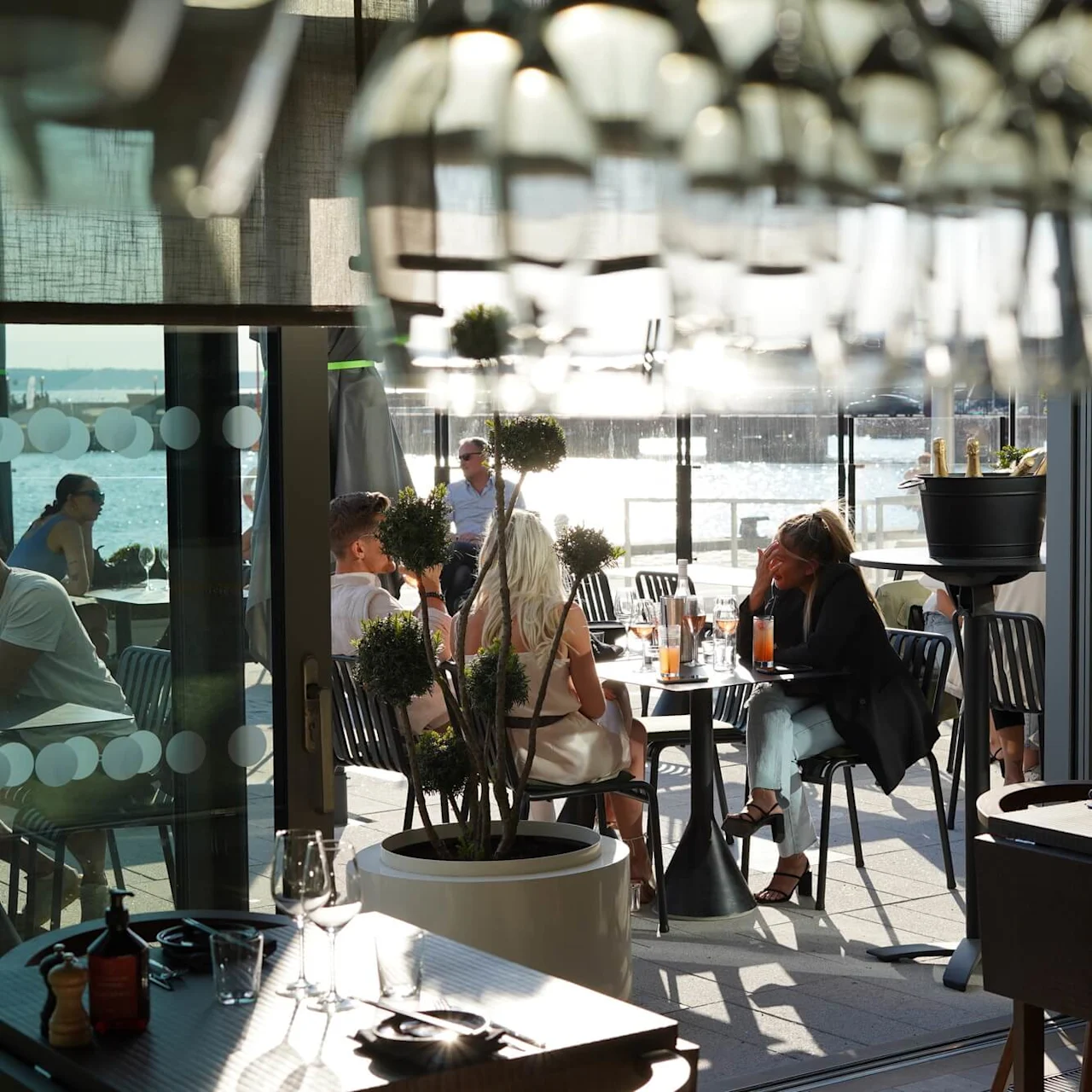 Folk sitter ved utendørs kafébord og nyter drikke under hengende glass, med sollys som reflekterer på vannet.