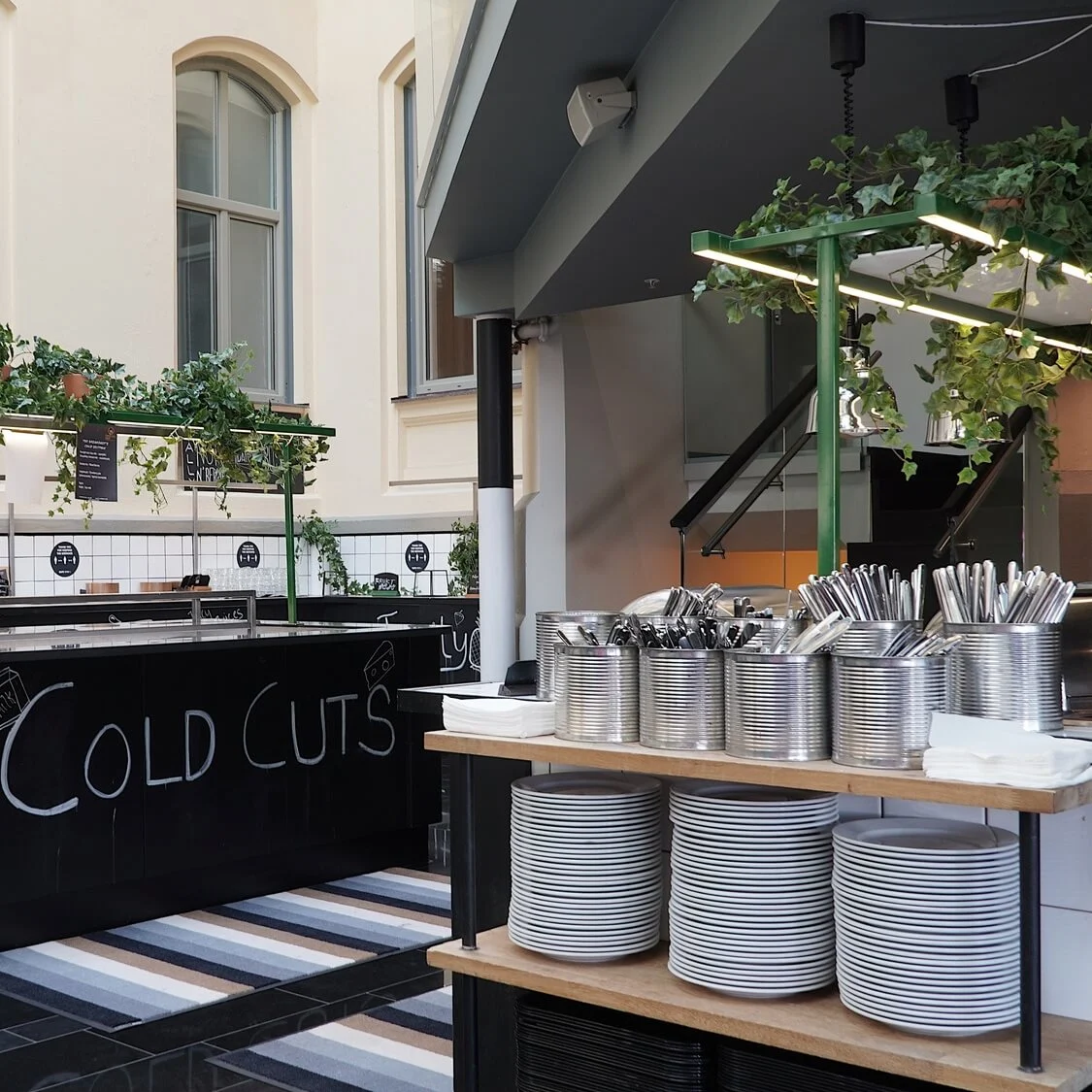 A buffet station with stacks of plates and cutlery, labeled "Cold Cuts," inside an airy, windowed space with black and white flooring.