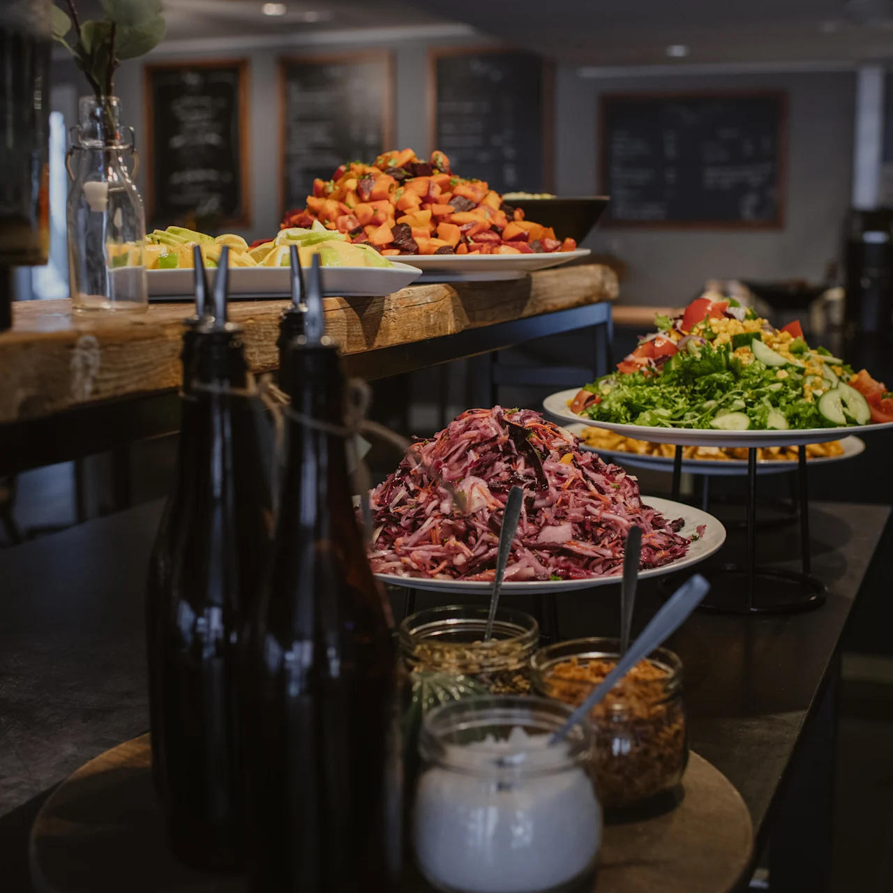 A buffet table with sliced meats, colorful salads, and condiments is displayed in a dining area with chalkboard menus on the walls in the background.