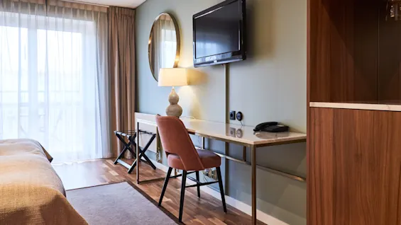 Hotel room with a bed on the left, work desk and chair in the center, and a wardrobe to the right, a TV mounted on the wall, and curtains drawn back to show daylight.