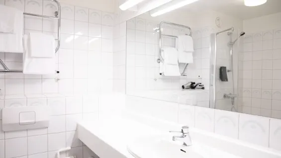 En hvid vask med en kromhane står på en stor bordplade; pænt foldede håndklæder hænger på stativet over, med en glasindelukket bruser synlig i det flisebelagte badeværelse.