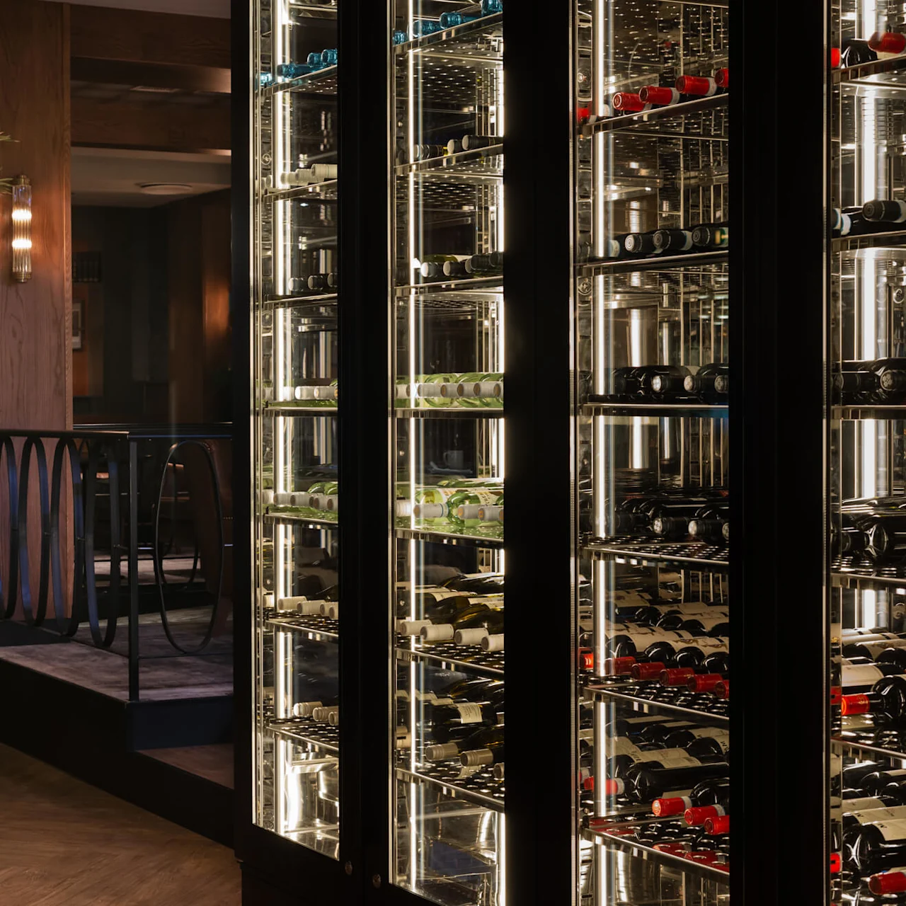 En vin-kylskåp visar upp olika flaskor på upplysta hyllor, intill en stor krukväxt i en dämpad restauranginteriör med trägolv.