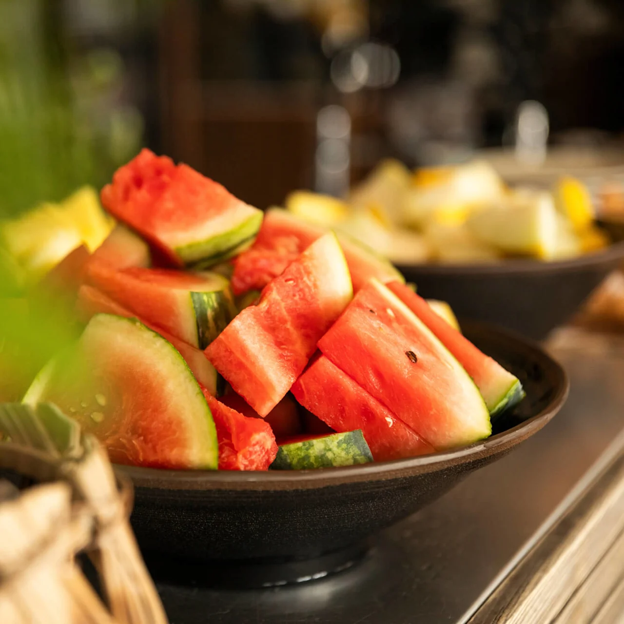Skivad vattenmelon fyller en mörk skål och står på ett bord. I bakgrunden syns fler fruktskivor, möjligtvis melon eller ananas, vilket förstärker en fräsch middagsupplevelse.