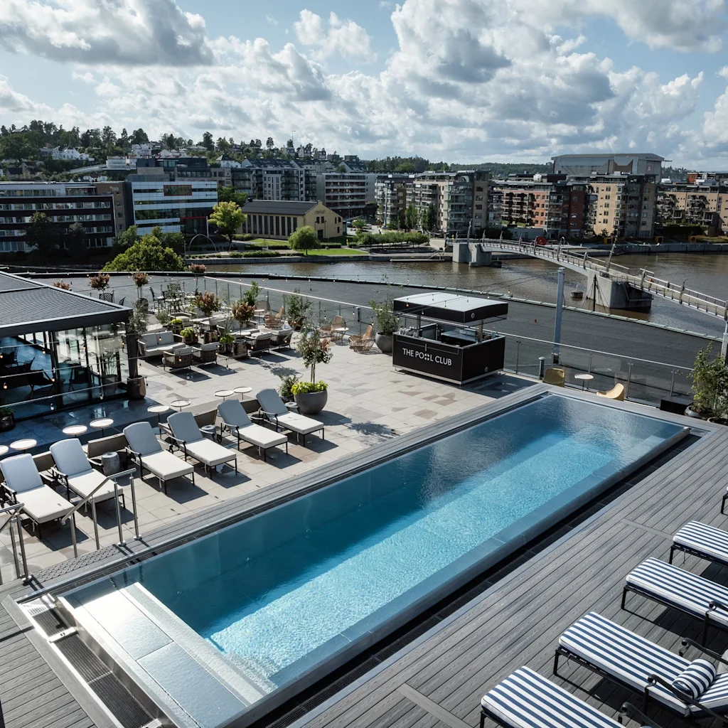 En tagpool med liggestole og potteplanter har udsigt over en flod og bybillede. "THE POOL CLUB" står på en nærliggende struktur.