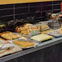 Bread and pastries are arranged on a buffet table, with baskets and trays, under tongs. Black tiled wall in the background reflects more pastries.