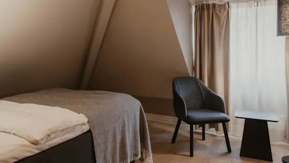 Et hyggeligt hotelværelse med seng, komfortabel stol og lille bord. Lyse gardiner ved vinduet skaber en fredelig atmosfære.