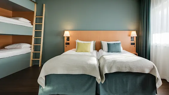 Two neatly made twin beds with pillows stand in a room with bunk beds and wooden flooring, illuminated by wall lamps, and surrounded by green walls and a window with curtains.
