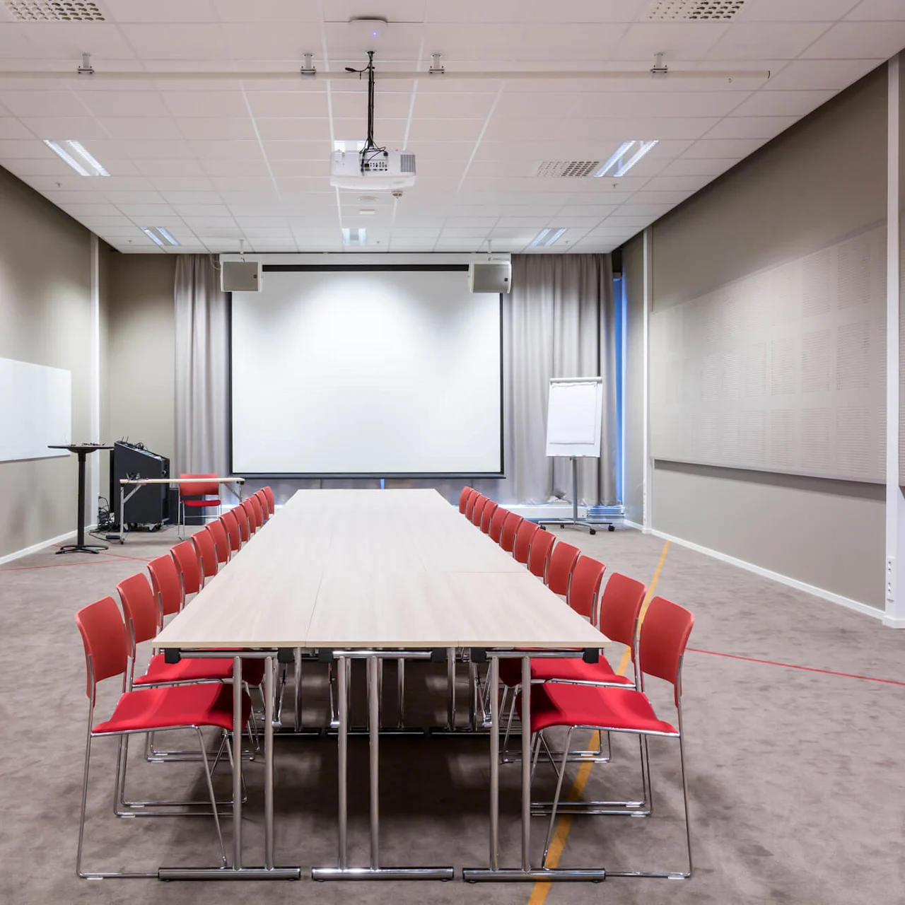 Ett långt konferensbord med röda stolar är centrerat i ett rymligt mötesrum. En projiceringsduk, takprojektor och blädderblock är synliga mot ljusa väggar.
