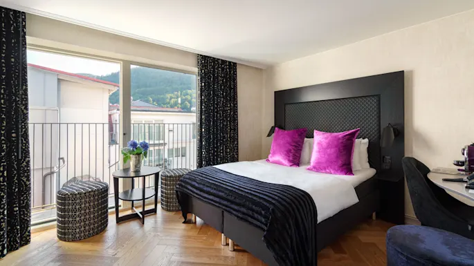 A bed with vibrant purple pillows is set in a modern room with wooden floors, beside a window overlooking buildings and greenery. A small table with flowers stands nearby.