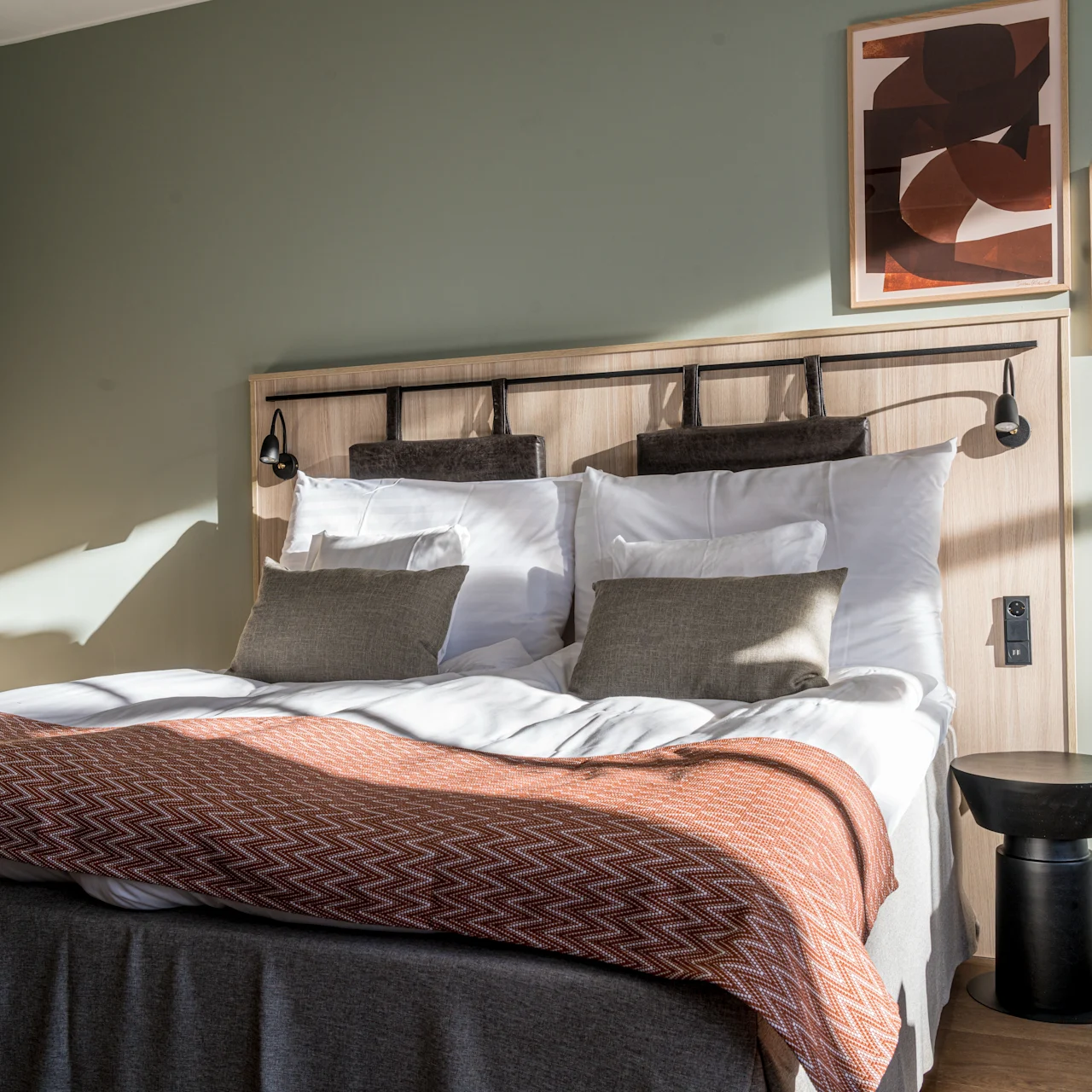 A neatly made bed features white pillows and a patterned brown blanket. It rests against a light green wall adorned with abstract art. Sunlight streams through a window.
