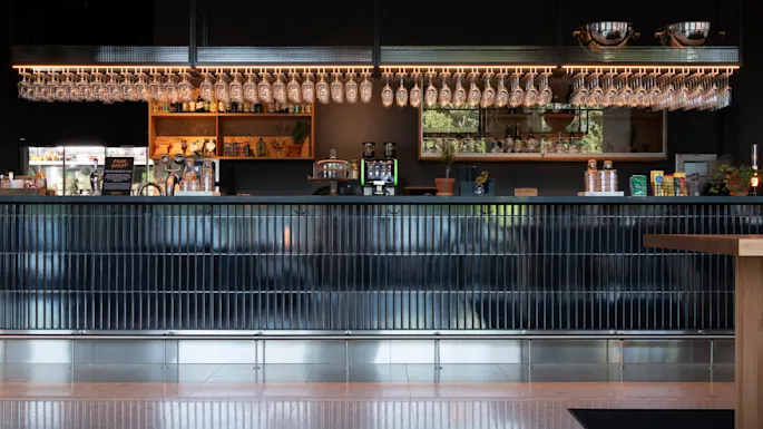 A sleek bar with hanging glasses, beverage taps, and a display of bottles set against a dark backdrop in a modern interior.