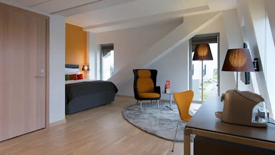 Ett modernt hotellrum har en säng och en mysig sittgrupp med en gul stol och svart fåtölj på en grå matta. Trägolv och vinklade tak ger unika designelement.