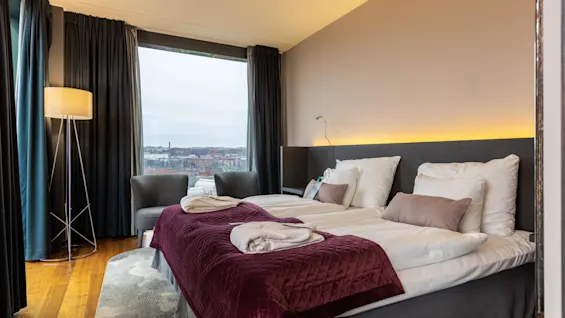 Two neatly arranged beds with burgundy quilts and pillows, situated in a modern room with a large window showing a cityscape. A floor lamp and armchair are nearby.