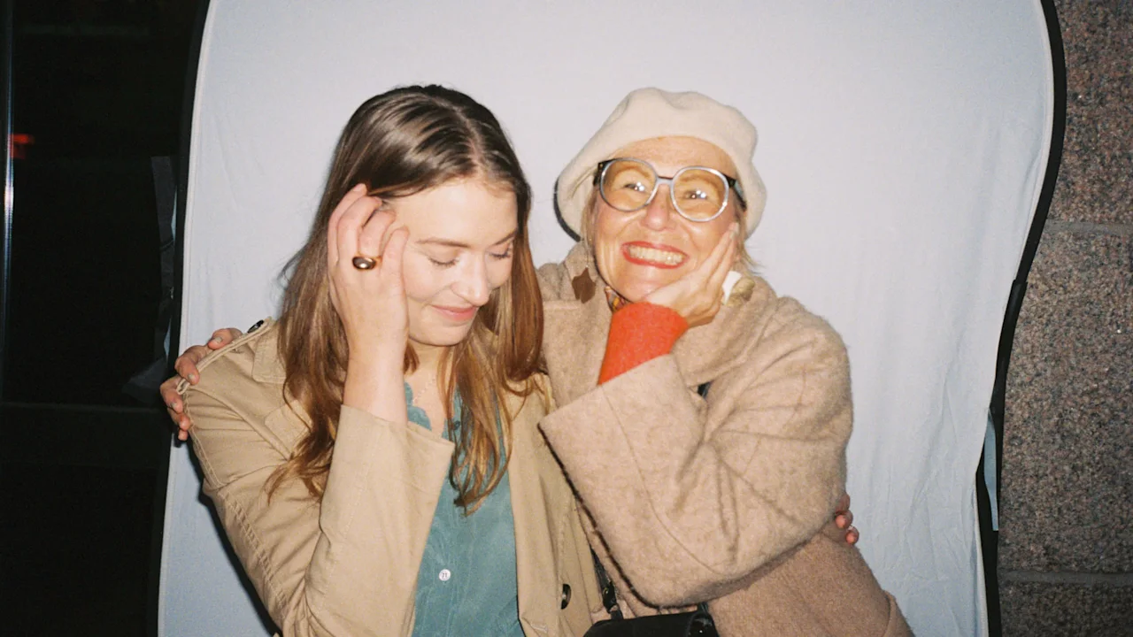 Two smiling women pose closely together. One covers her face shyly while the other, wearing glasses and a hat, hugs her. They stand in front of a plain backdrop.