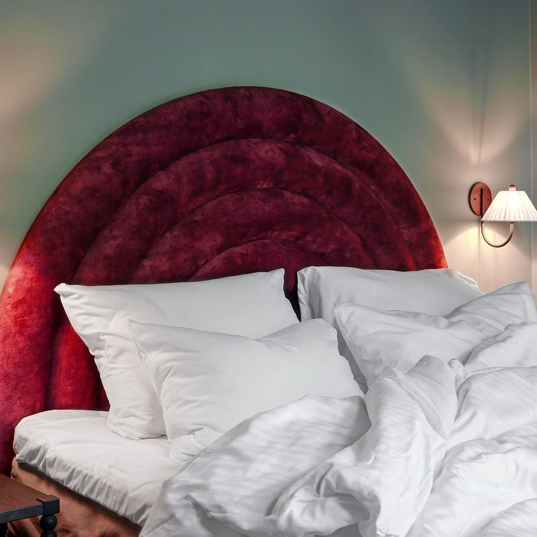 A bed with white rumpled sheets and pillows sits beneath a red, plush headboard. Wall-mounted lamps illuminate the room, with a nightstand holding a book and a vase of flowers.