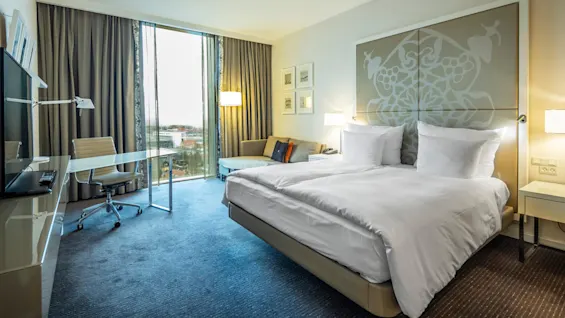 Ett väl upplyst hotellrum med en stor säng, dekorativ sänggavel och sänglampor. Ett skrivbord med en stol är bredvid ett fönster med gardiner, bredvid en soffa och golvlampa.