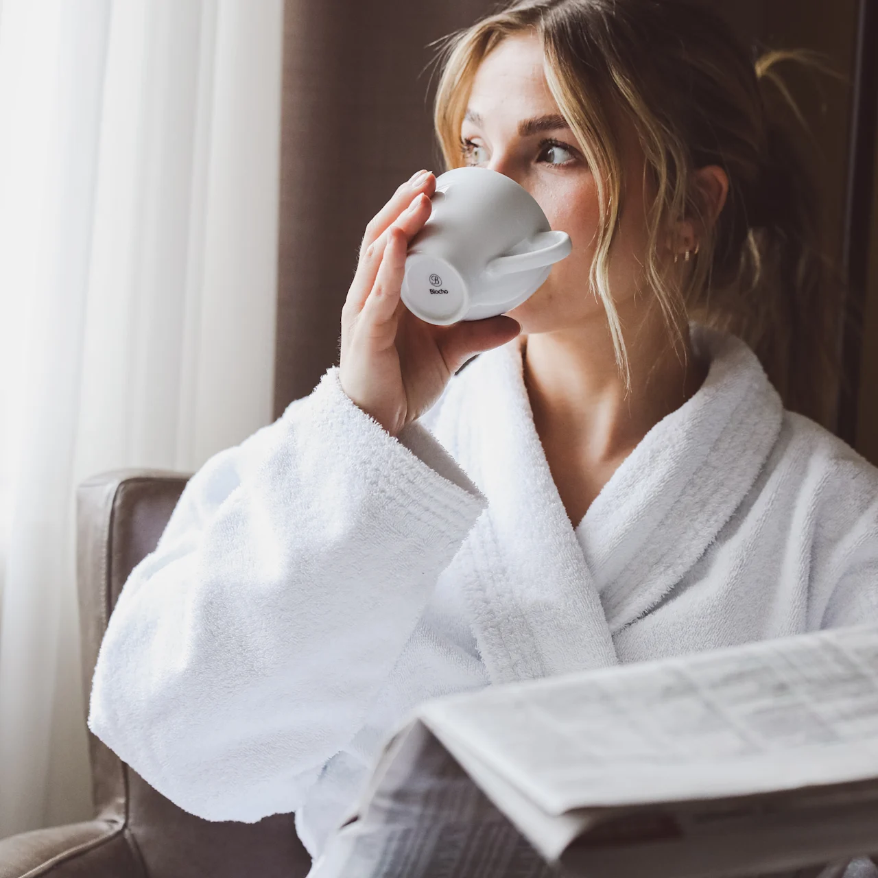 A person in a white robe sits by a window, sipping from a mug and holding a newspaper. The room features soft lighting and a fabric curtain.