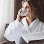 A person in a white robe sits by a window, sipping from a mug and holding a newspaper. The room features soft lighting and a fabric curtain.