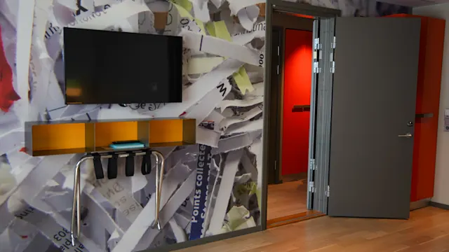 En väggmonterad TV på en tapet med ett mönster av sönderrivet papper, ovanför en hylla med prydligt vikta föremål. En öppen dörr avslöjar en ljusröd korridor.
