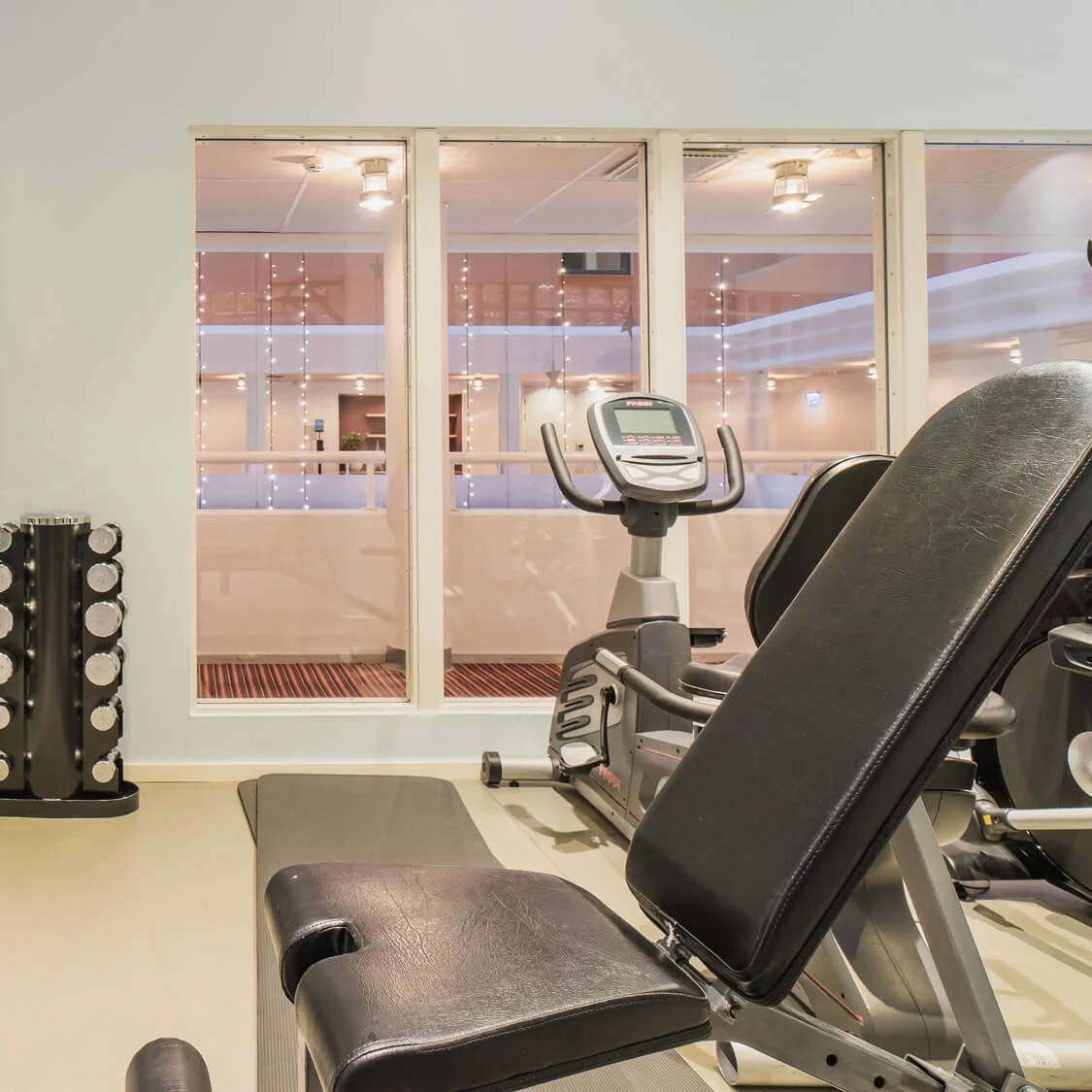 Exercise bench sits unused beside mirrored wall; nearby exercise bikes rest under fluorescent lights with windows revealing empty corridor. Dumbbells neatly stacked between bench and bikes.