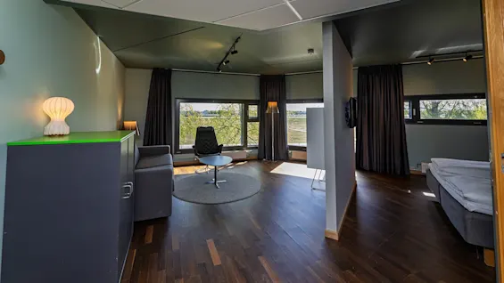 A modern room with a black swivel chair on a rug, facing large windows. A gray sofa, bed, cabinets, and curtains complement the space, illuminated by lamps.