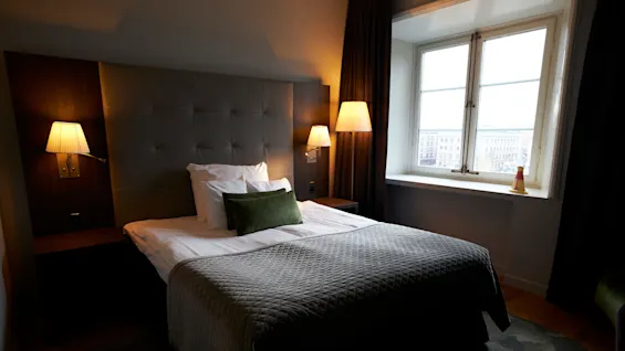 A neatly made bed with a green decorative pillow and quilted blanket is illuminated by two bedside lamps in a cozy, softly lit hotel room with a large window.