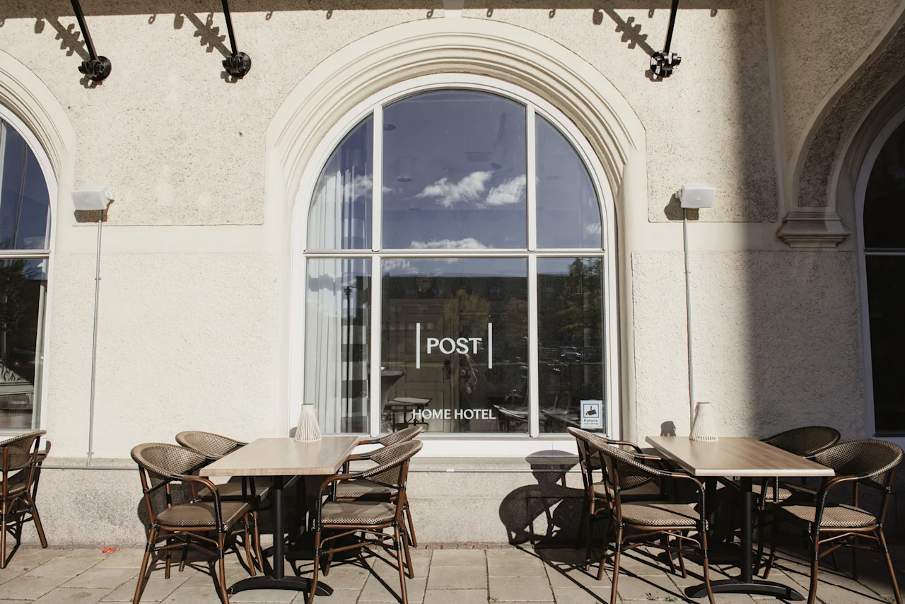 Uteservering framför en byggnad med ett stort välvt fönster. Fönstret visar texten POST HOME HOTEL.