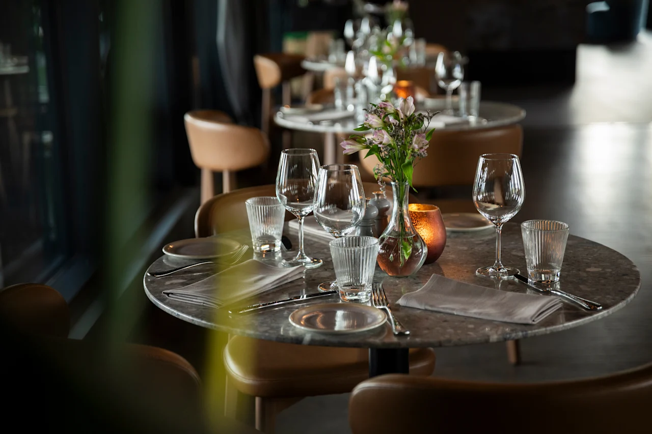Et elegant dekket restaurantbord med vinglass, vannglass, bestikk, servietter, blomster og et varmt lys.