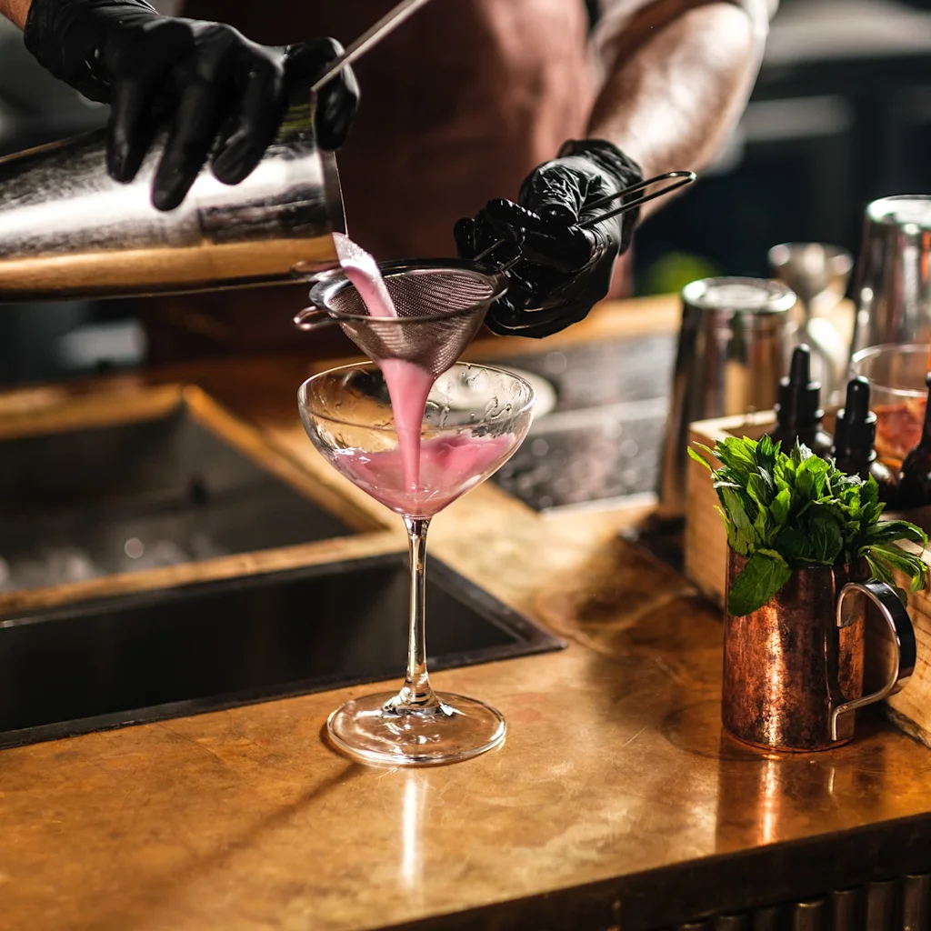 En bartender i svarte hansker siler forsiktig en livlig rosa cocktail i et martiniglass på en varm trebar.