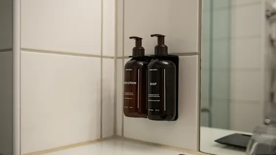 Two wall-mounted brown bottles labeled "LOTION" and "SOAP" with pumps, fixed on a white tiled bathroom wall near a mirror and sink.