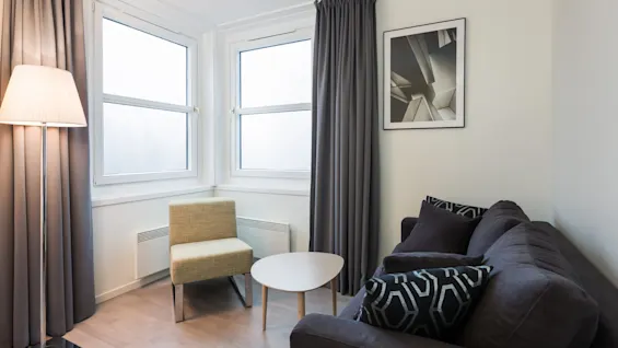 A gray sofa, featuring patterned cushions, sits beside a round table in a modern living room. A nearby lamp illuminates the corner, with large windows and a geometric artwork on the wall.