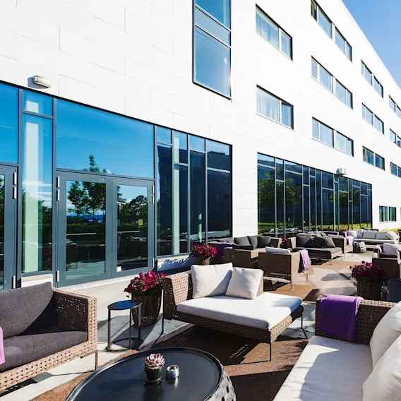 Utendørssitteplasser har flettede sofaer og bord arrangert på en uteplass. Plasseringen skaper en avslappende atmosfære rundt den moderne bygningens reflekterende vinduer, under klar blå himmel og frodig grønt.