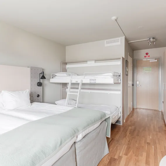 Valoisa hotellihuone, jossa kaksi erillistä sänkyä ja kerrossänky, puulattia ja ulos johtava ovi.