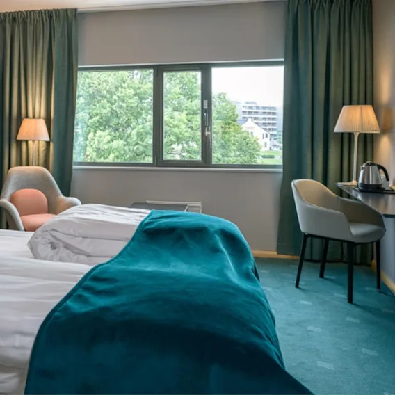 Et hotellrom med pent oppredd seng, fremhevet med teal puter og et kast, et sitteområde og utsikt over grøntareal fra vinduet.