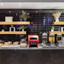 A breakfast buffet features pastries and beverages on a counter with black tiles and wooden shelves adorned with plants and decorative items.