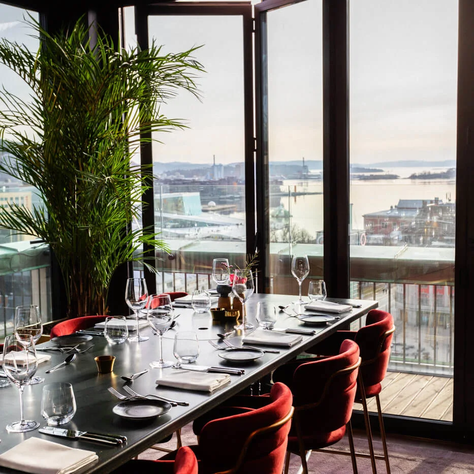 A dining table set with red chairs, glasses, and napkins. The room features large windows overlooking a harbor with sunlight streaming, and a tall plant nearby.