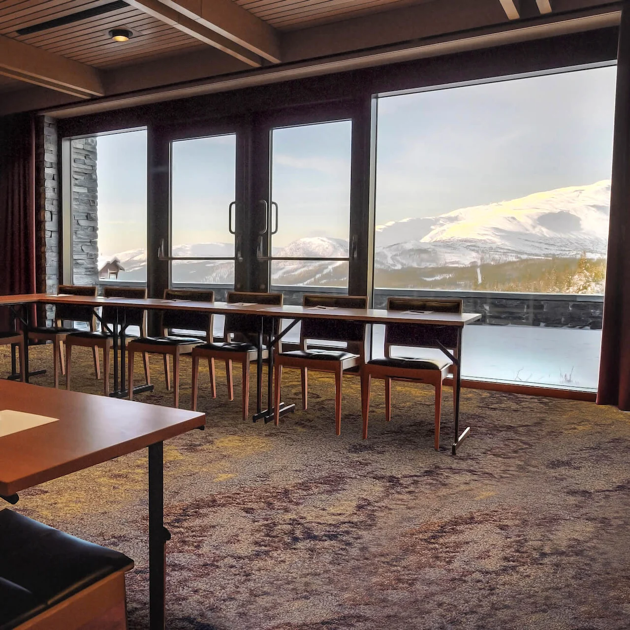 Meeting room with arranged tables and chairs, set with notepads and pens, facing large windows. Snow-covered mountains are visible through the windows, providing a scenic backdrop. 