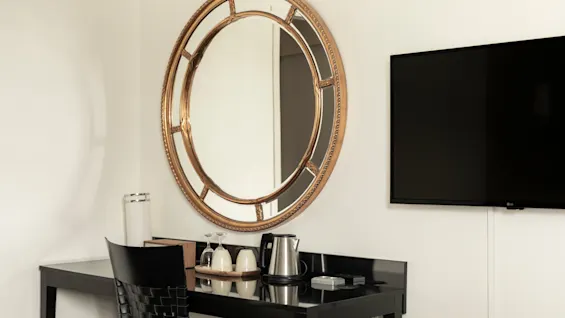 Hotellihuoneen työpöytäalue, jossa on koristeellinen pyöreä peili, seinälle asennettu televisio ja vedenkeitin kuppeineen juomia varten.