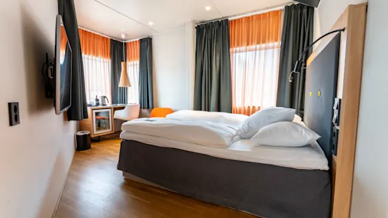 En seng med hvidt sengetøj er placeret i et hotelsværelse. Den vender mod et fladskærms-TV på væggen. Store vinduer med orange gardiner lyser det trægulvede rum op.