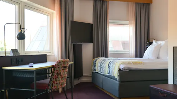 En pænt oppredt seng står i et hyggeligt, godt oplyst hotelværelse med to vinduer, et vægmonteret TV, et skrivebord med lampe og en mønstret stol.