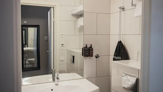 A white bathroom sink reflects in a mirror beside a towel rack with folded towels and dark bottles. A wall-mounted toilet and a black bag labeled "A-D-W-E-R" complete the scene.