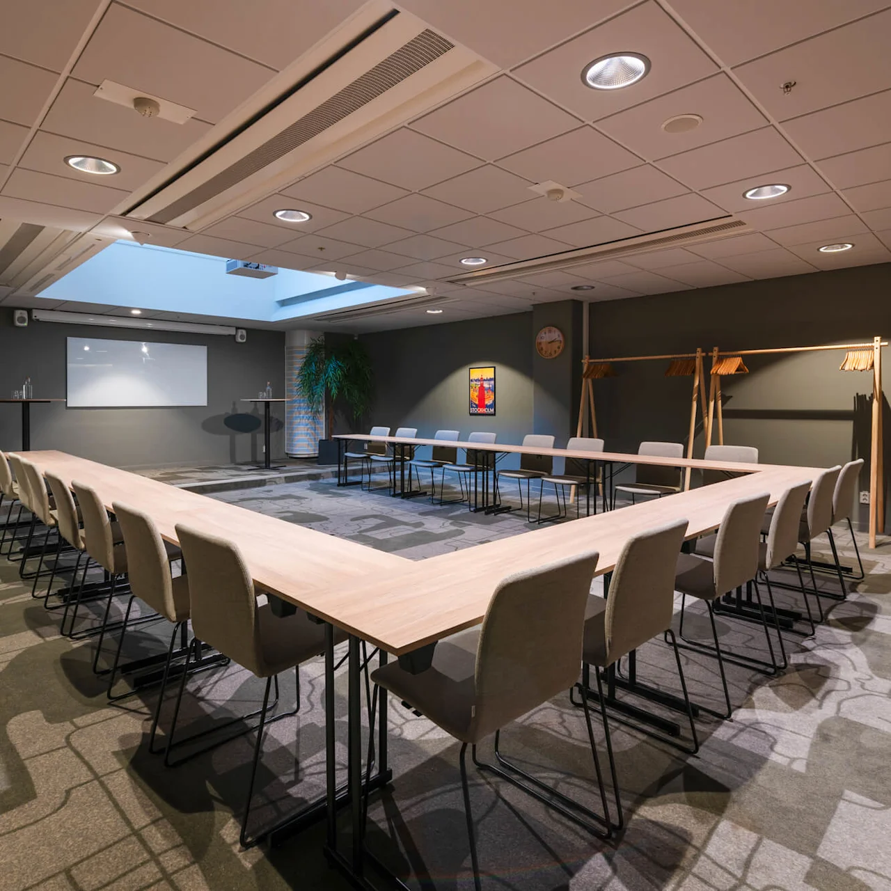 U-shaped conference table with beige chairs surrounds a boardroom, featuring a projector screen, plants, and abstract carpet. A clock and art piece are on the wall.