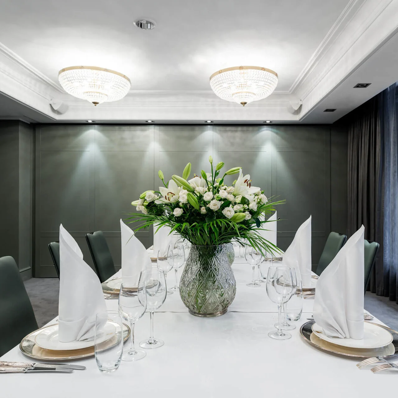 A table adorned with an elegant floral arrangement in a vase, surrounded by neatly arranged plates, glasses, and folded napkins in a sophisticated dining room with chandeliers and grey walls.